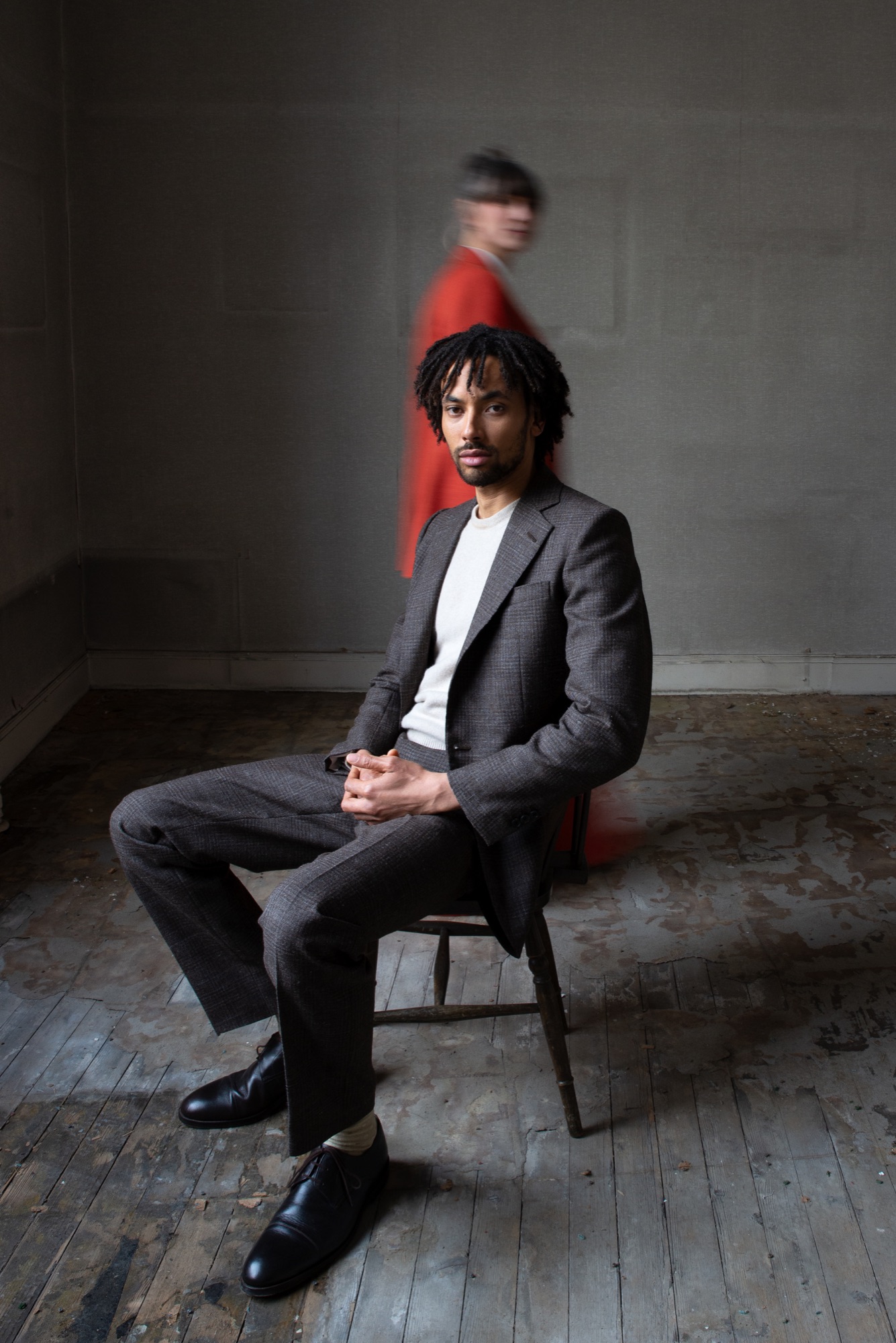 A man seated in a hand-tailored grey wool suit, a figure in red passing behind him
